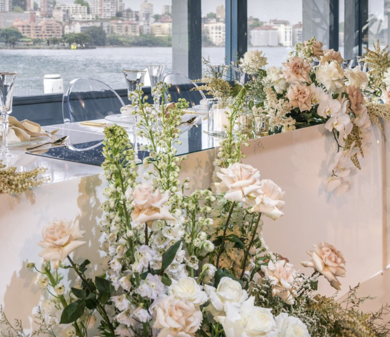 Elegant dining table setup with floral centerpiece near windows overlooking city and water view.