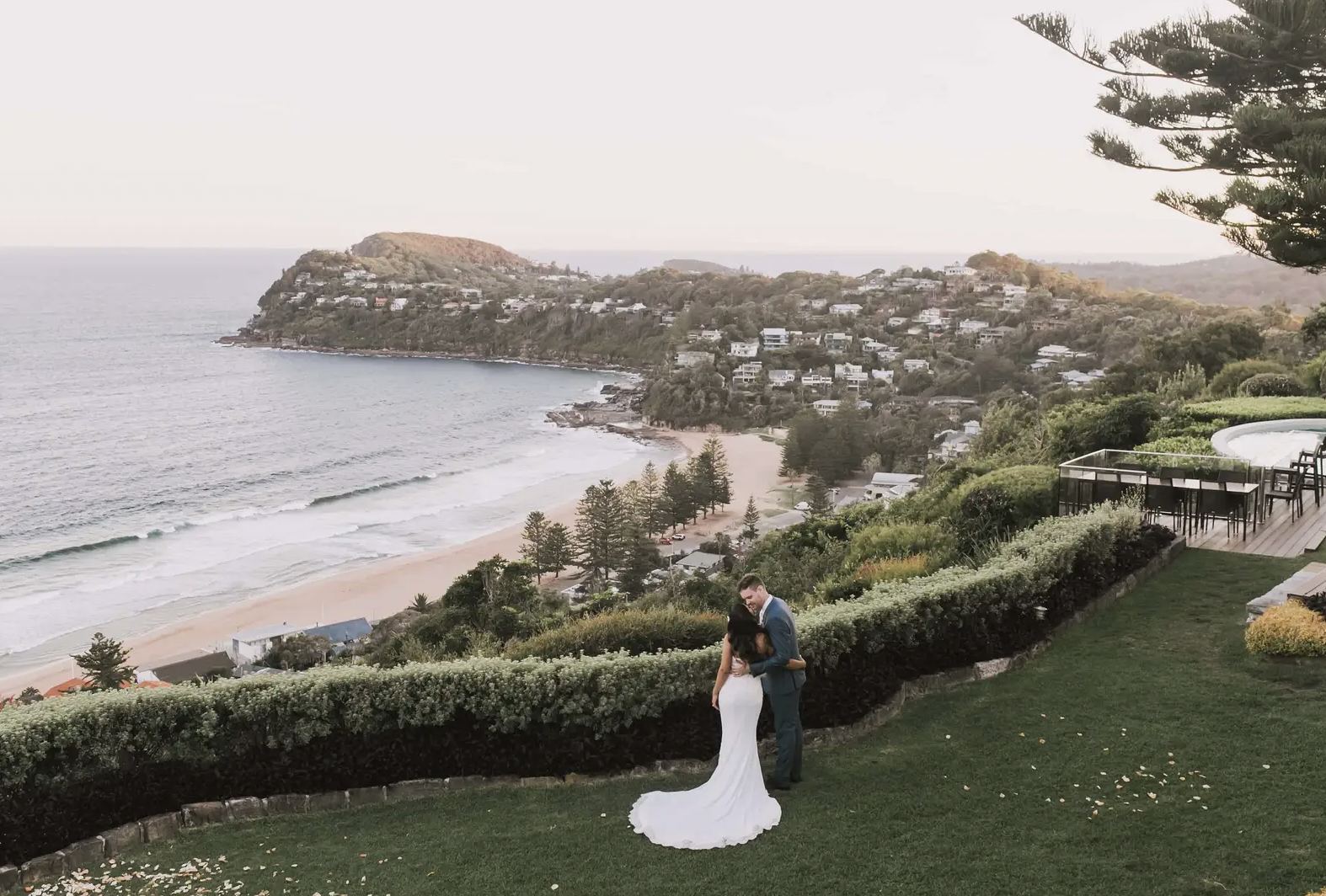 A bride and groom embracing on a grassy hill overlooking a beach and hillside neighborhood, with trees and a large pine tree on the right side.