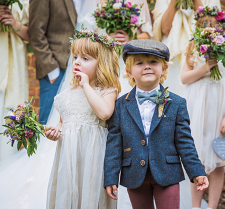 Children at a wedding, a girl with a flower crown and a dress, and a boy with a hat and suit, standing and holding flowers, surrounded by other kids in dresses and suits holding bouquets.