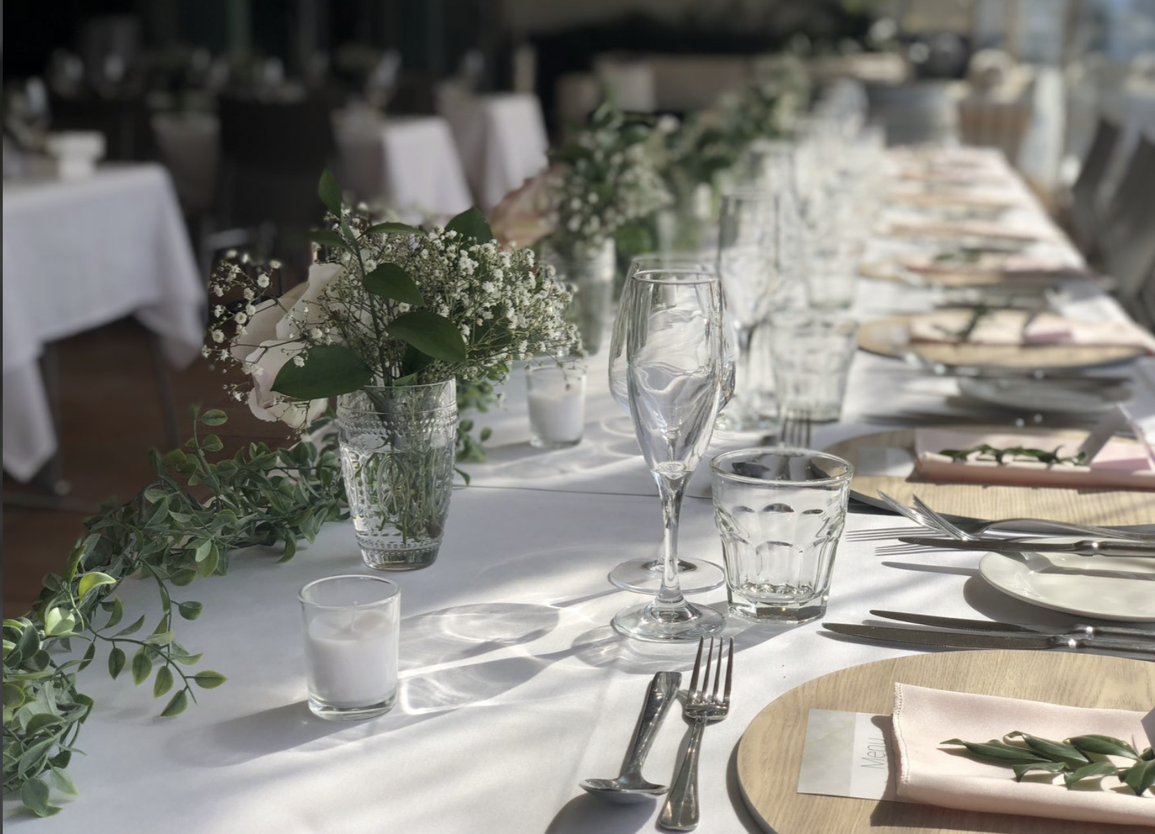 A long dining table set for a wedding with white tablecloths, floral centerpieces, and place settings including plates, napkins, glasses, and cutlery. The table is decorated with green foliage and white flowers, illuminated by natural light.