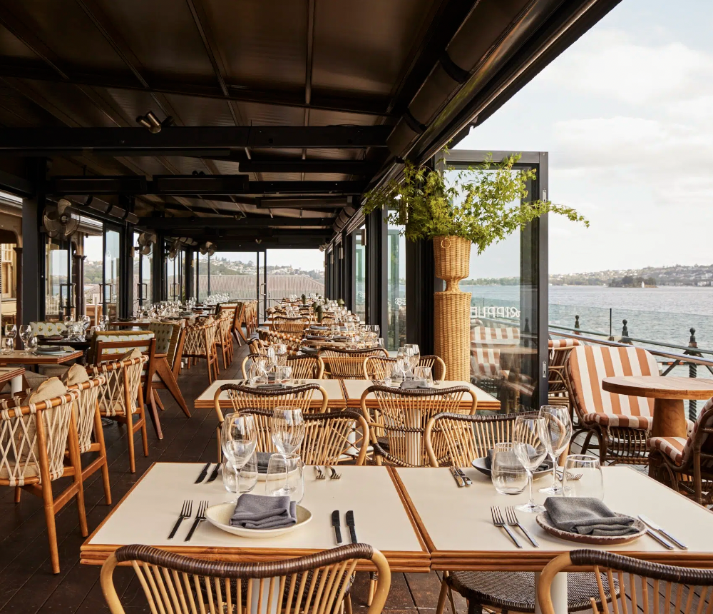 A spacious, covered outdoor restaurant dining area with tables set for meals, overlooking a body of water with a distant shoreline, decorated with potted greenery and striped cushions on outdoor seating.