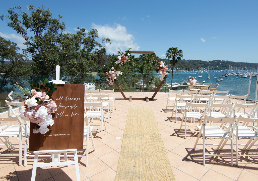 Outdoor wedding ceremony setup at a seaside location with chairs, a floral arch, and a welcoming sign, overlooking a marina with sailboats and palm trees under a clear blue sky.