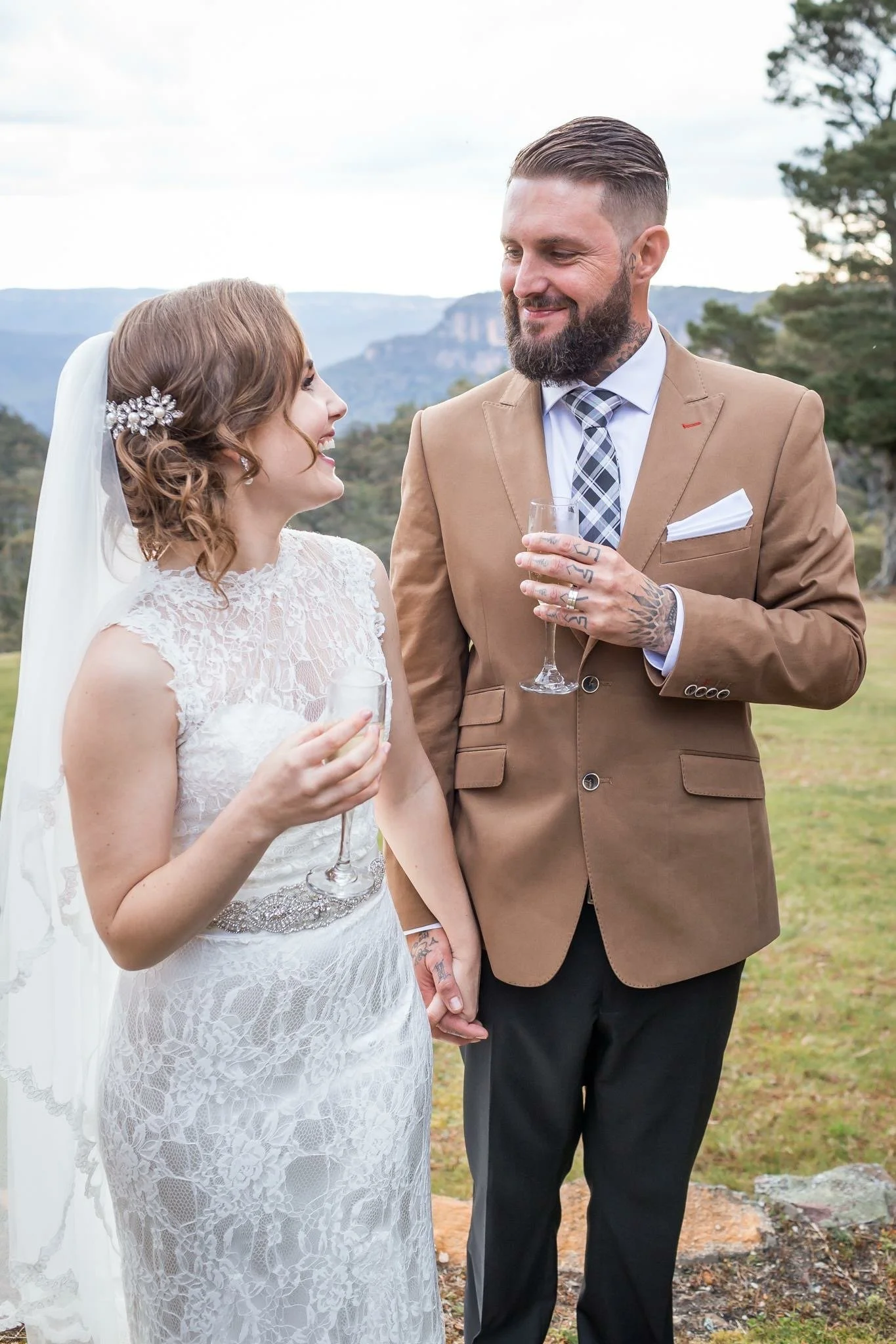 A bride and groom holding hands and looking at each other during their wedding outdoors. The bride is wearing a white lace wedding dress with a veil and decorative hairpiece. The groom is in a beige suit with a plaid tie and has tattoos on his hands and neck. They are smiling and holding champagne glasses. In the background, there are trees and mountains.