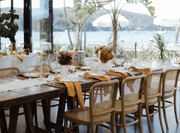 Set dining table with white and rust-colored napkins, vases with floral arrangements, and candles in a restaurant overlooking a body of water and mountains.
