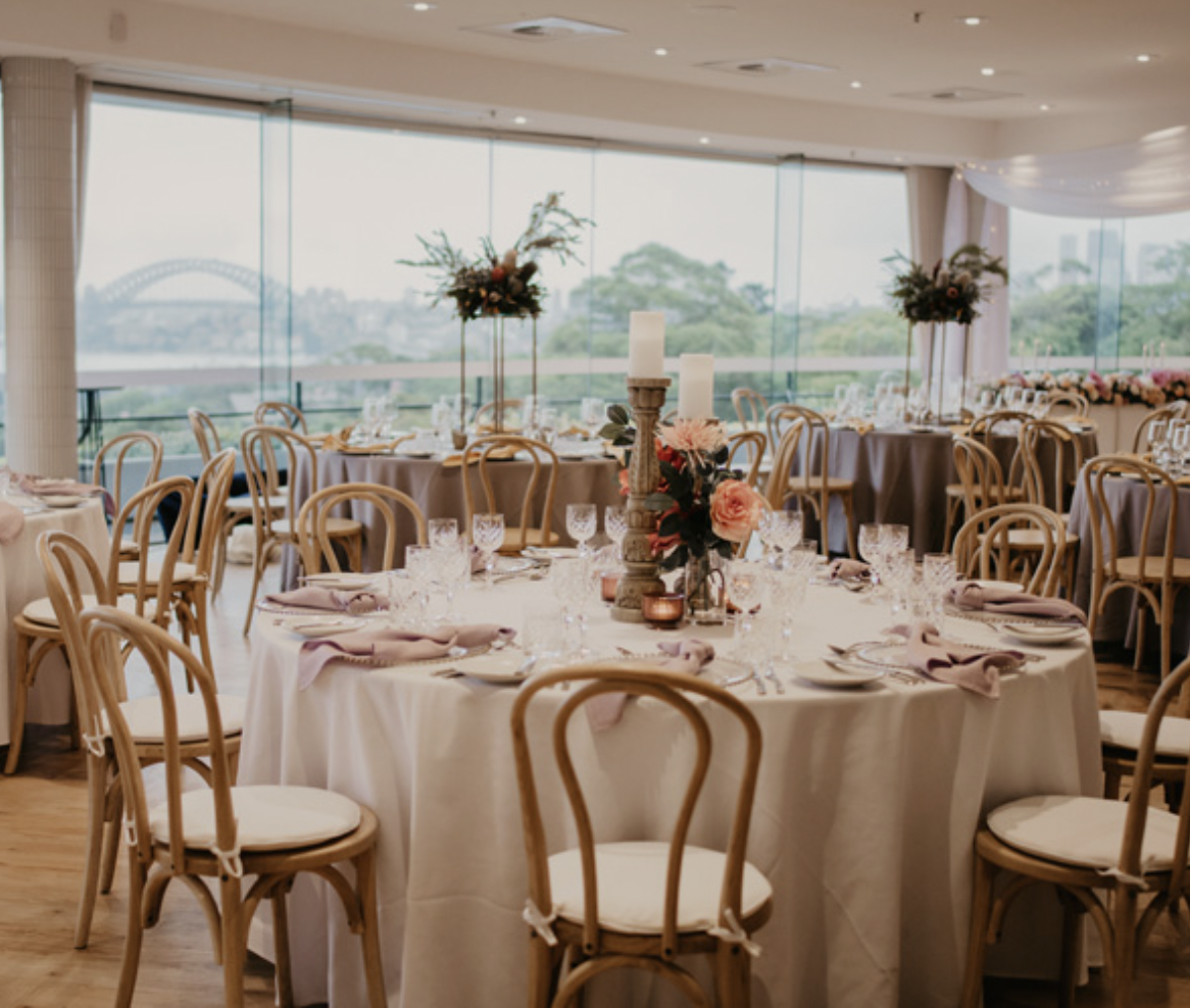Elegant banquet hall with round tables decorated with pink napkins, floral centerpieces, candles, and fine glassware. Large windows reveal a cityscape with water and a bridge in the background.