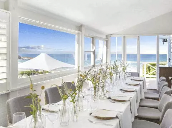 A long dining table set for a meal with white tablecloths, plates, glasses, and small flower arrangements, overlooking the ocean through large windows.