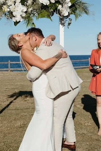 A bride and groom sharing a kiss during their outdoor wedding ceremony by the ocean, with an officiant standing nearby.