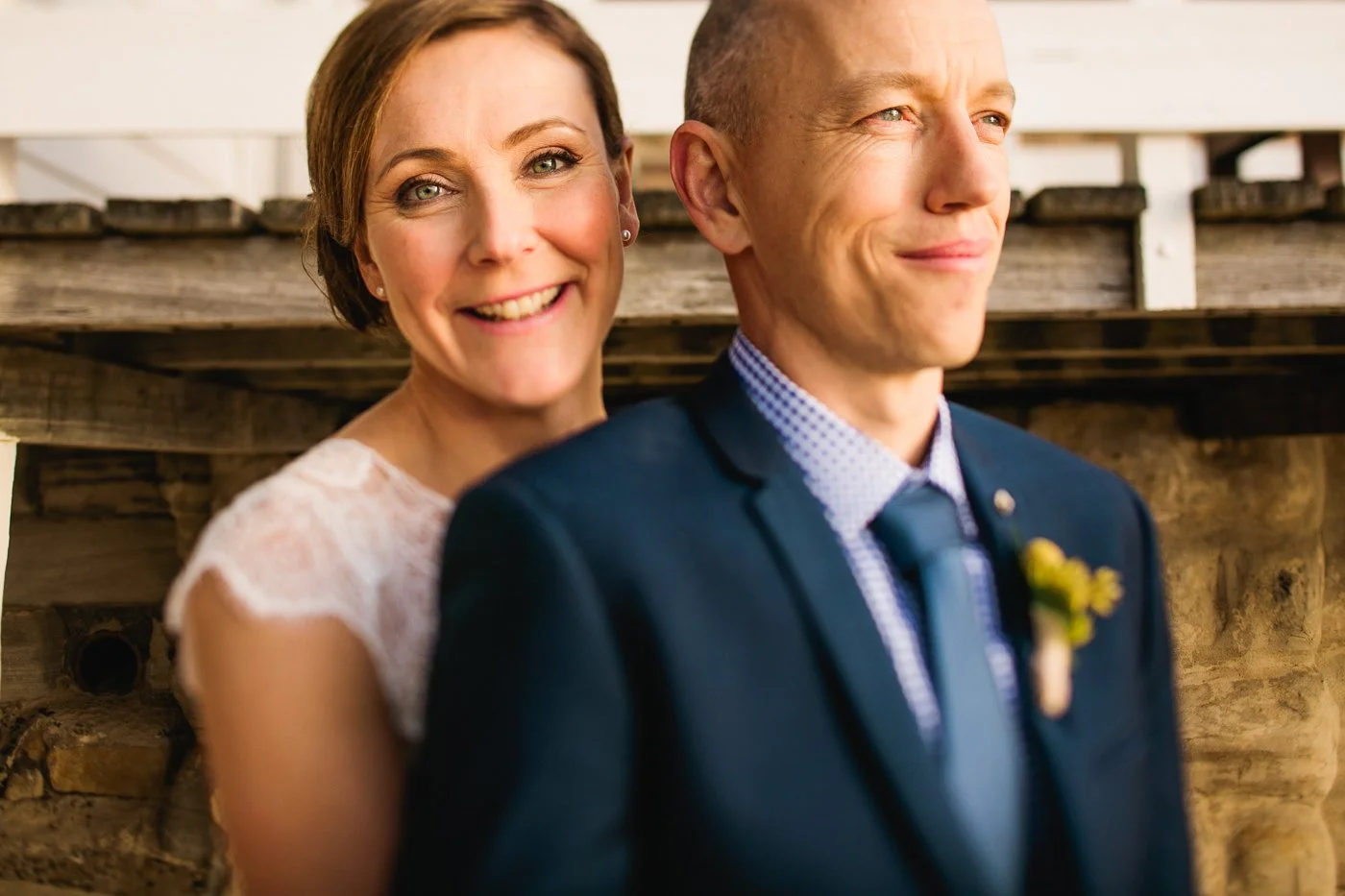 A smiling woman with short brown hair and earrings standing close behind a man in a navy suit, matching shirt, and tie, wearing a boutonniere, against a rustic outdoor background.