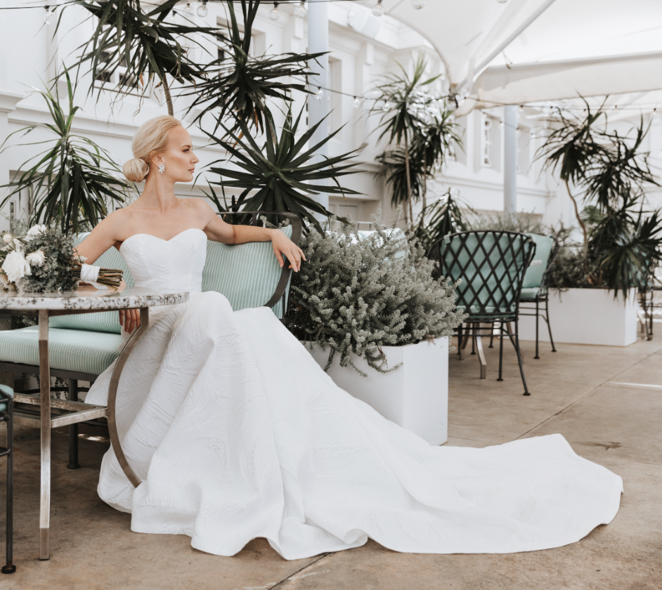 A woman in a white wedding dress sitting on a light green bench in a sunlit conservatory or greenhouse surrounded by large potted plants and greenery.