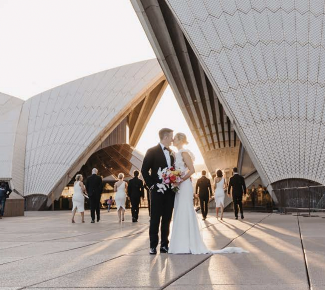 A bride and groom at their wedding, kissing in front of the Sydney Opera House during sunset. Wedding guests are walking in the background.