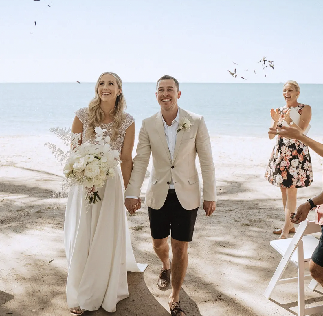 A couple getting married on the beach, holding hands and smiling, with friends celebrating nearby.