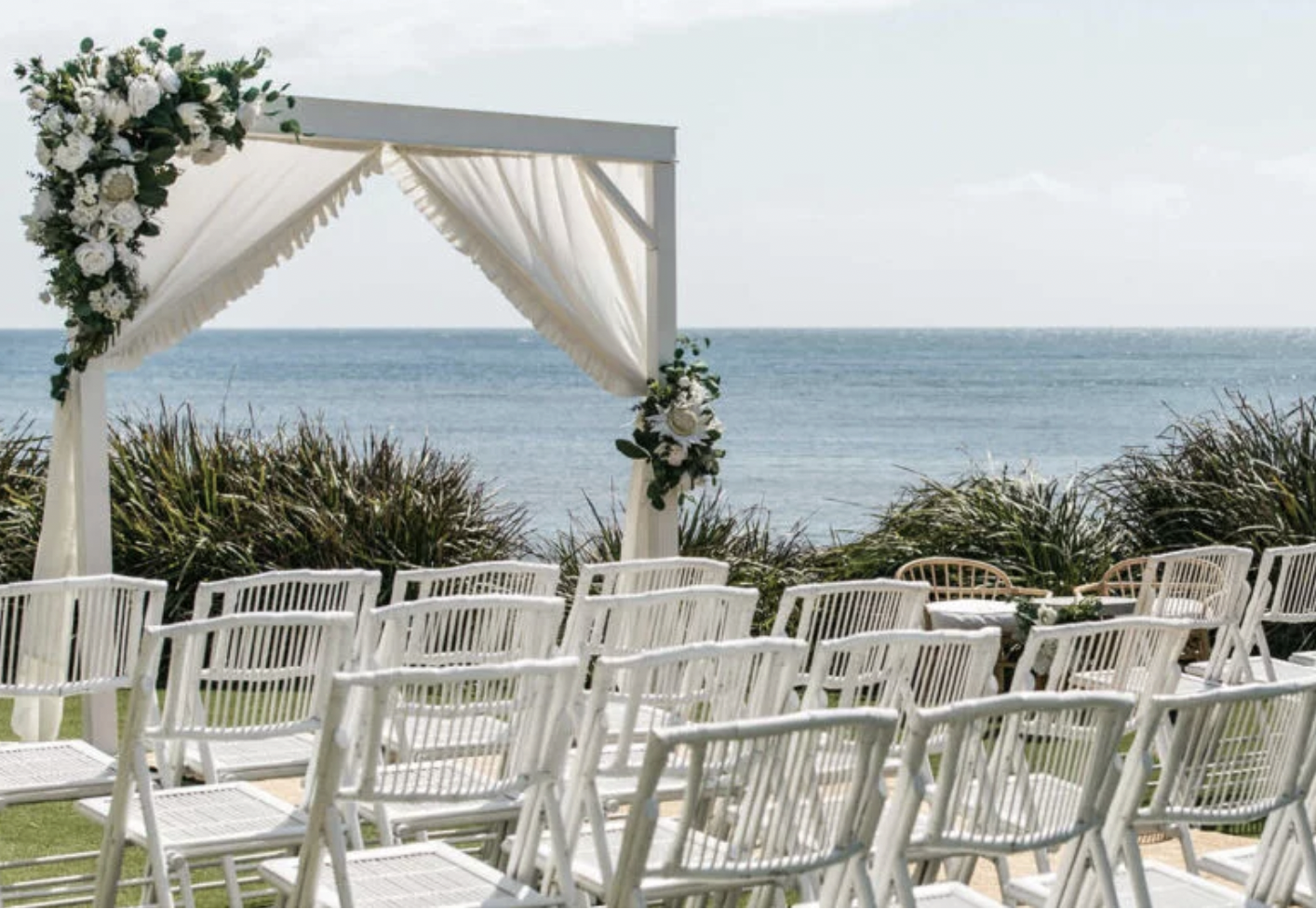 Outdoor wedding ceremony setup with white chairs under a decorated canopy on a grassy area overlooking the ocean and sky.