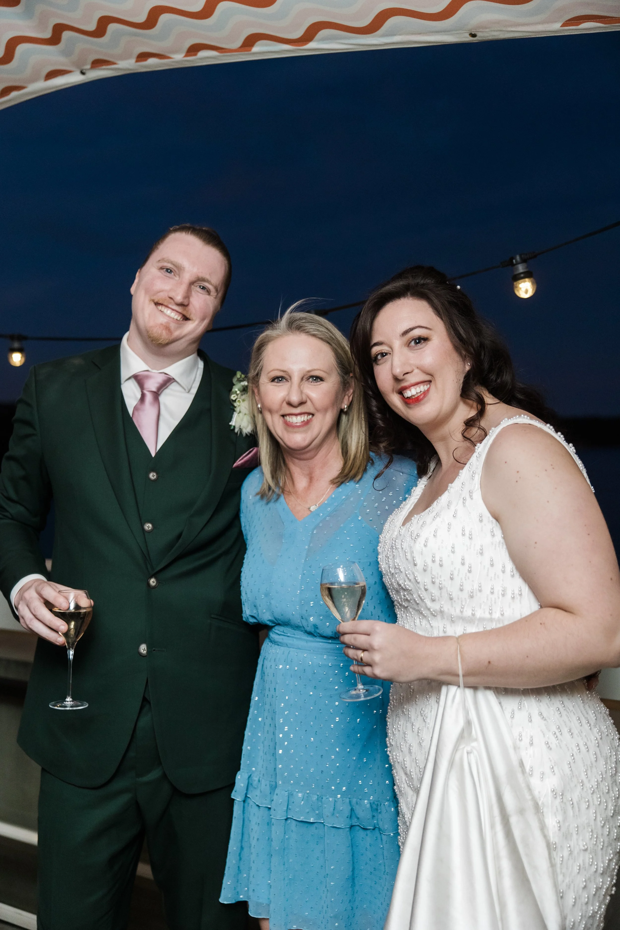 Three adults at a celebration, smiling and holding glasses of champagne, standing under string lights in an outdoor setting at night.
