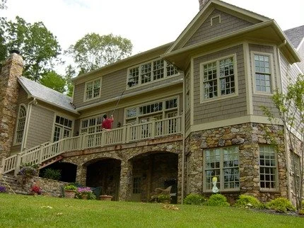 A large multi-story house with a stone foundation and wooden siding, featuring multiple windows, a balcony, and a staircase leading to the yard, surrounded by greenery and trees.