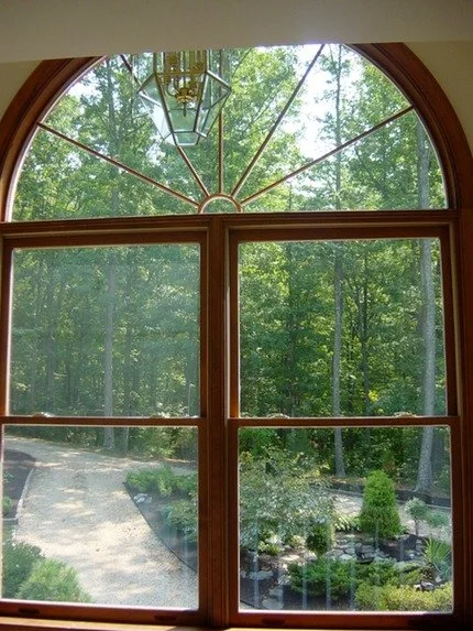 View of a lush green wooded area through a large arched window with a wooden frame, featuring a geometric metal decorative element and a hanging lantern in the upper part of the window.