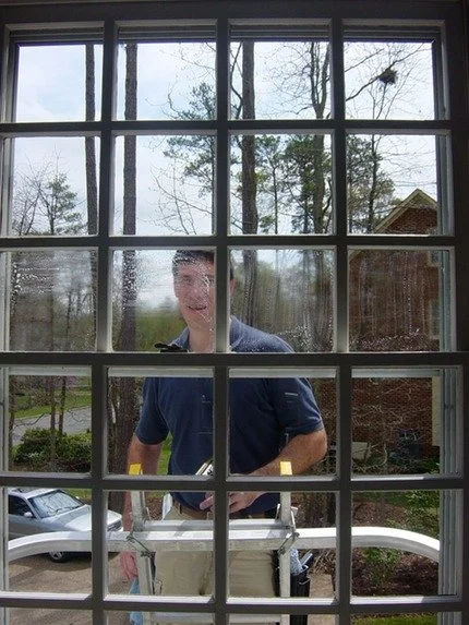 A young man standing outside and smiling, viewed through a multi-pane window, with a street, parked car, trees, and a house in the background.