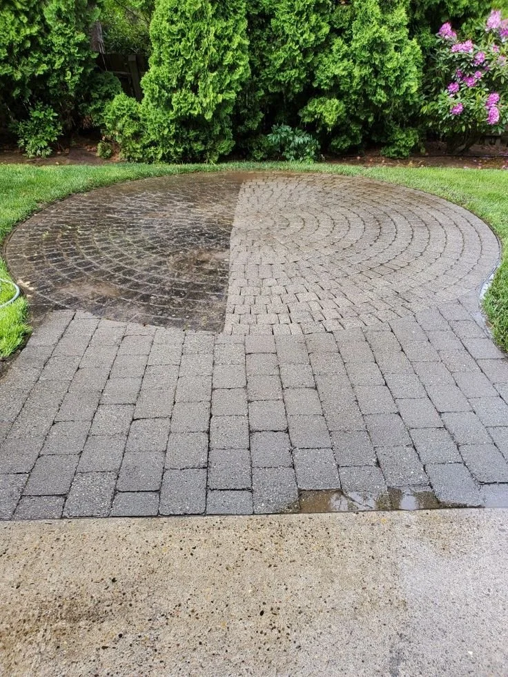 A circular brick patio area with a side by side comparison of the same area before and after cleaning, bordered by green grass and bushes with pink flowers in the background.