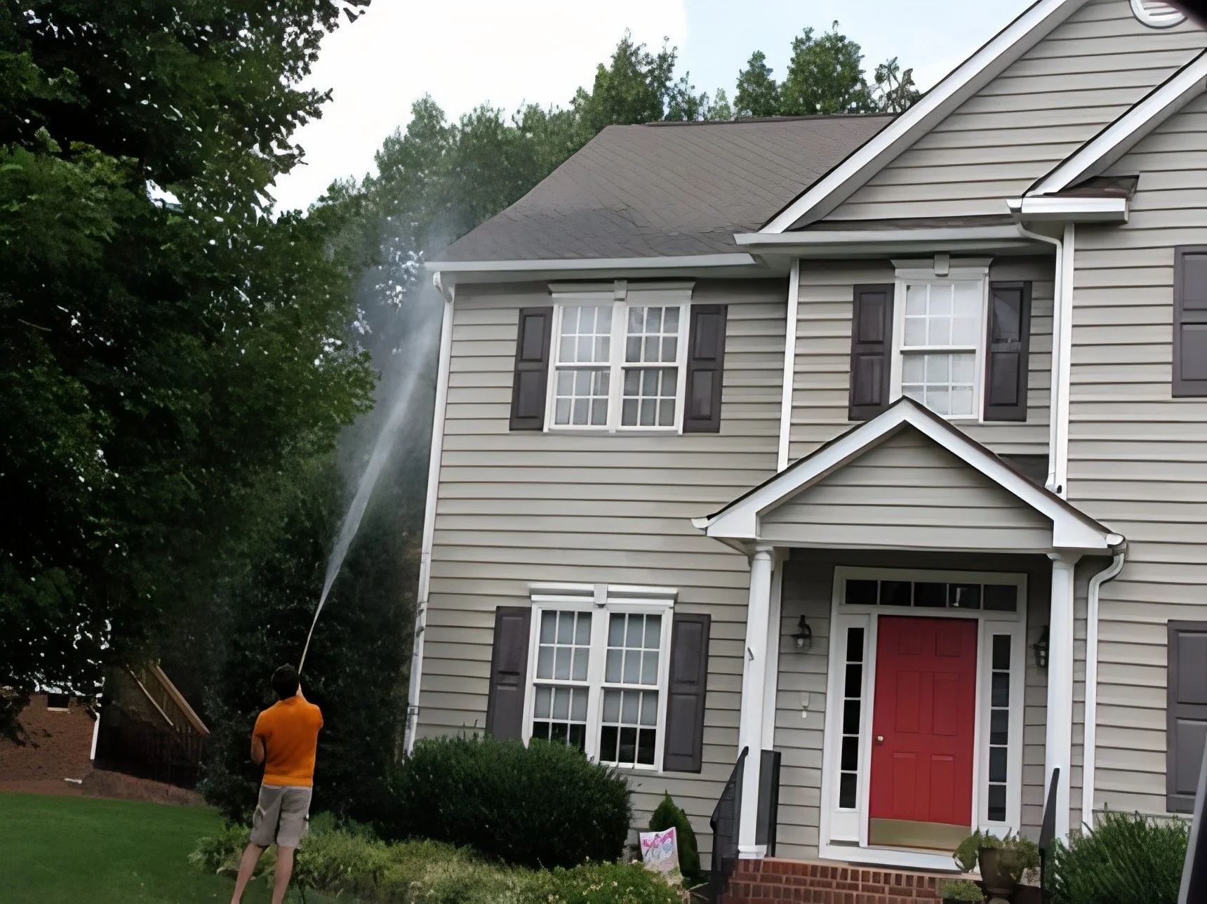 A person in an orange shirt and khaki shorts using a pressure washer to clean the side of a grey house with white trim and black shutters. The front door is red, and there are trees and a lawn in the background.