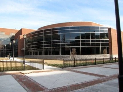 Modern multi-story office building with glass windows and brick exterior, surrounded by a walkway and a black fence.