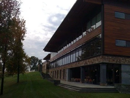 A modern multi-story building with large glass windows and wooden exterior, surrounded by a grassy area and trees, under cloudy skies.
