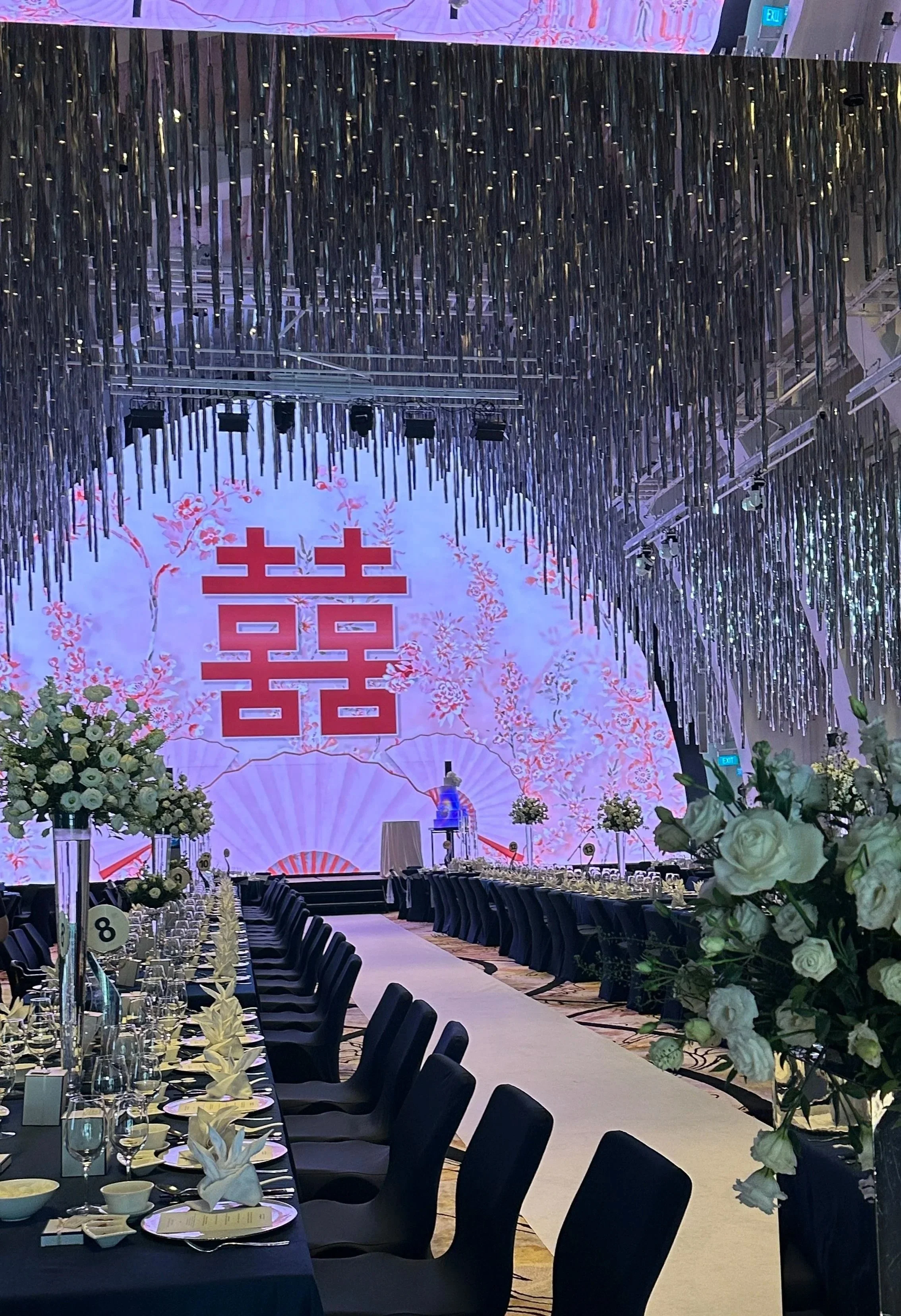 Elegantly decorated banquet hall with long dining tables set with glassware, plates, and napkins, floral centerpieces, floral backdrop with a large double happiness symbol, hanging metallic streamers, floral arrangements in the foreground, and stage 