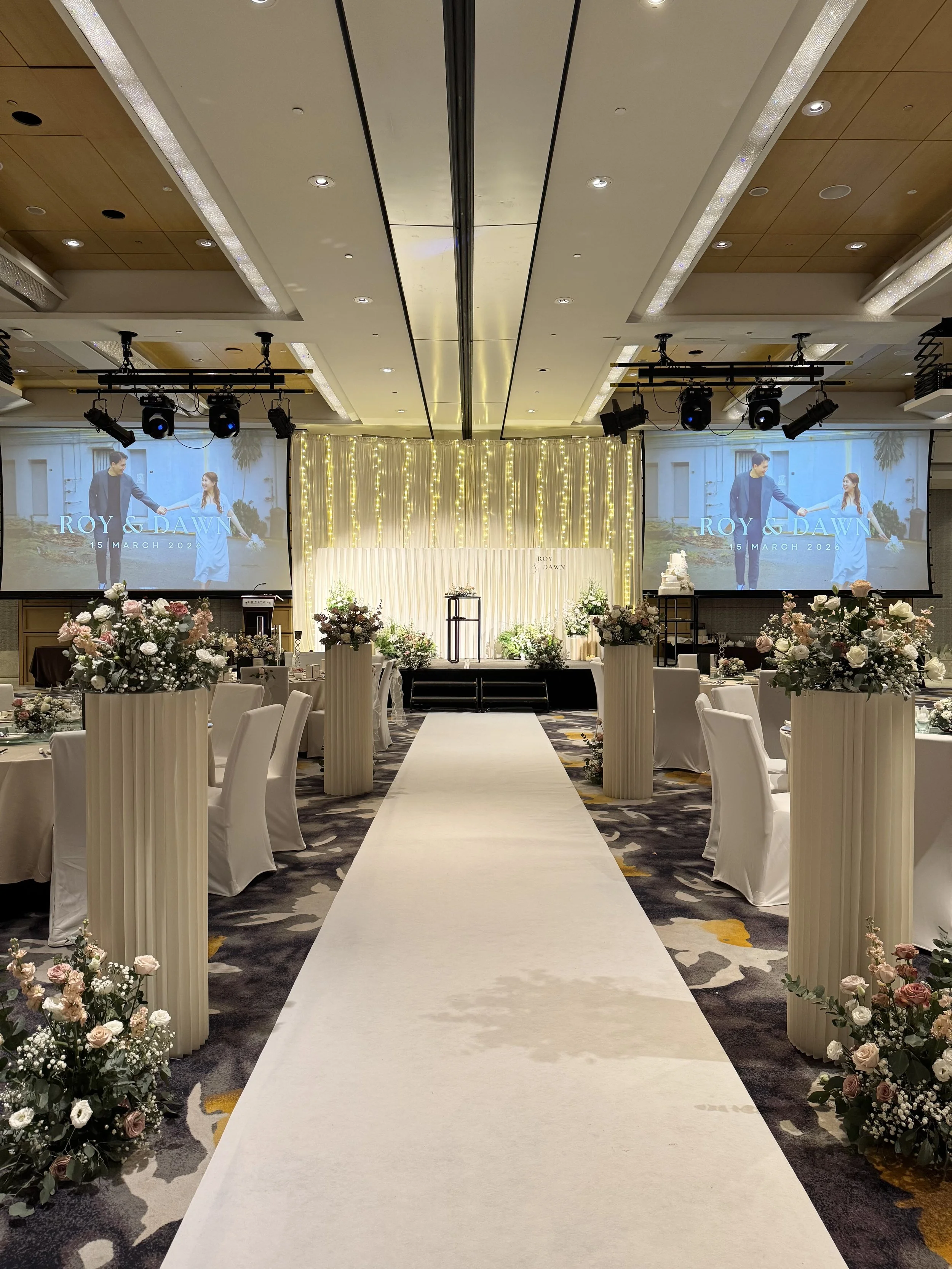 Elegant wedding ceremony setup with a white aisle runner, floral arrangements, and a decorated stage backdrop with string lights, two large screens displaying the names 'Roy & Dawn' and the date '15 March 2026'.