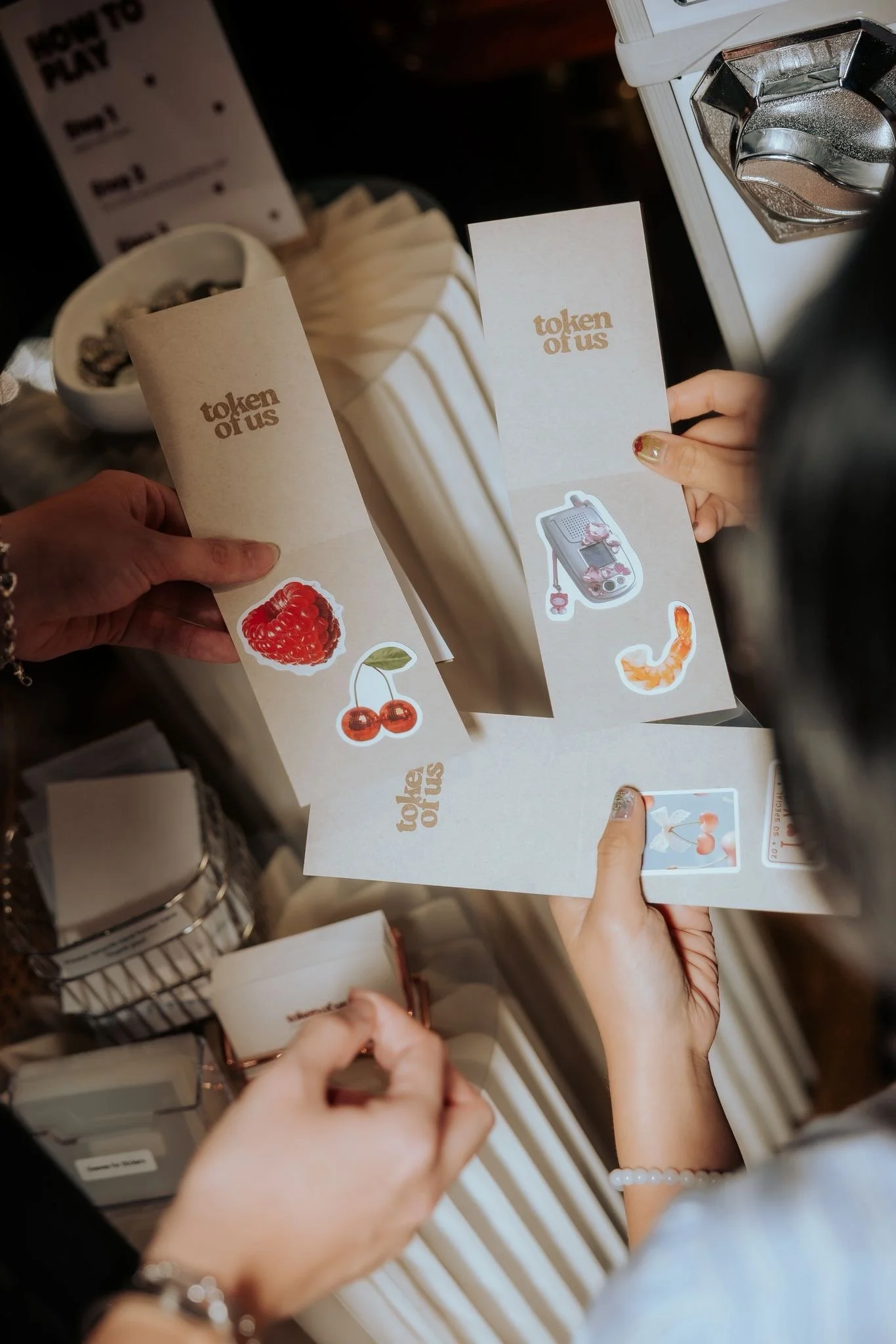 Multiple hands holding beige paper inserts with the text "token of us" in a retail or store setting. Background includes a bowl of coffee beans, decorative items, and a white object with a metallic dial.