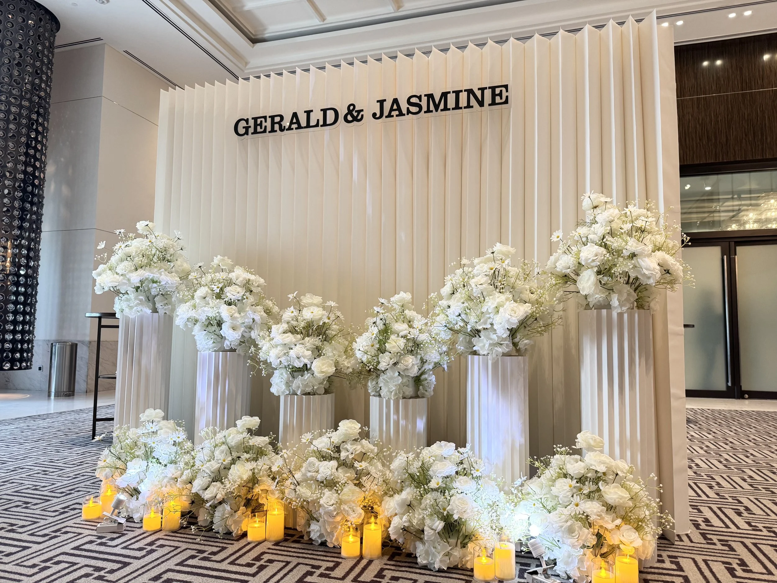 A wedding or event decoration with a backdrop that has the names "Gerald & Jasmine". The backdrop is cream-colored with vertical pleats. In front, there is a floral arrangement with white flowers and greenery, illuminated by candles placed on the flo