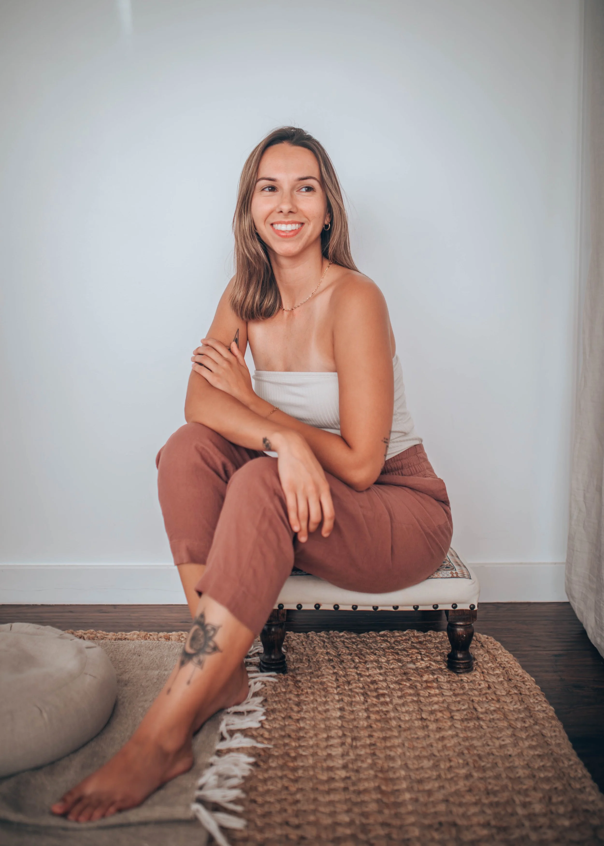 A woman sitting on a cushioned bench with a smile, looking to her left, wearing a white strapless top and brown pants, her arms crossed over her knees, with a tattoo on her left ankle and another on her wrist. The room has a beige rug and a simple wall background.