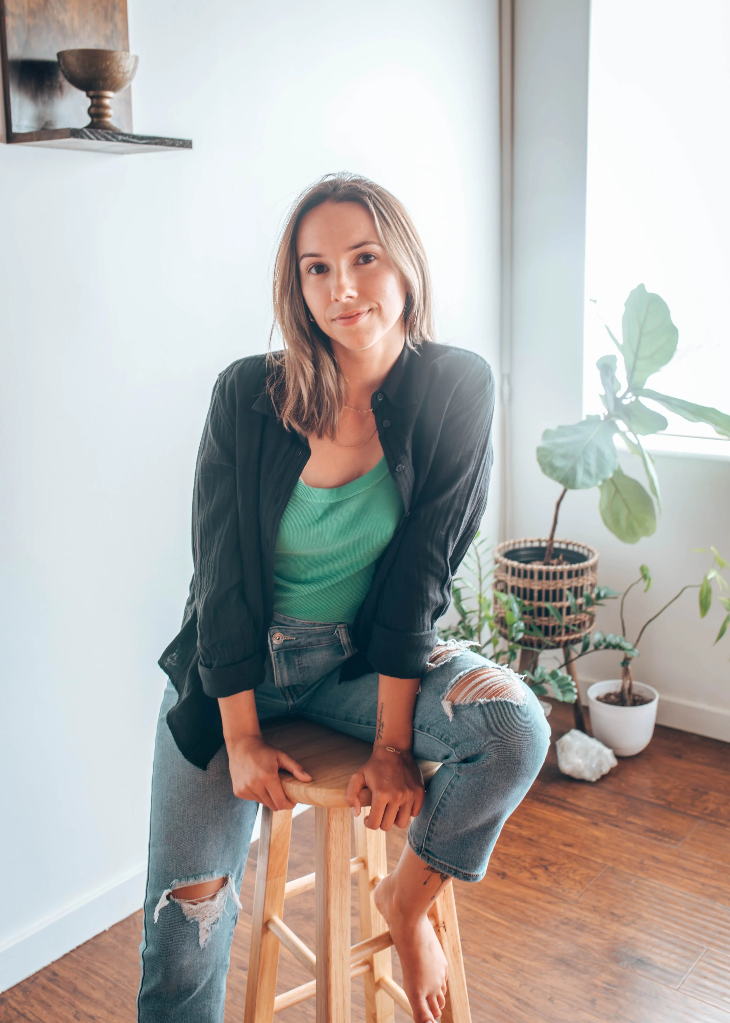 A young woman with shoulder-length brown hair, wearing a black shirt over a green top and ripped jeans, sits on a wooden stool in a well-lit room with hardwood floors and potted plants.