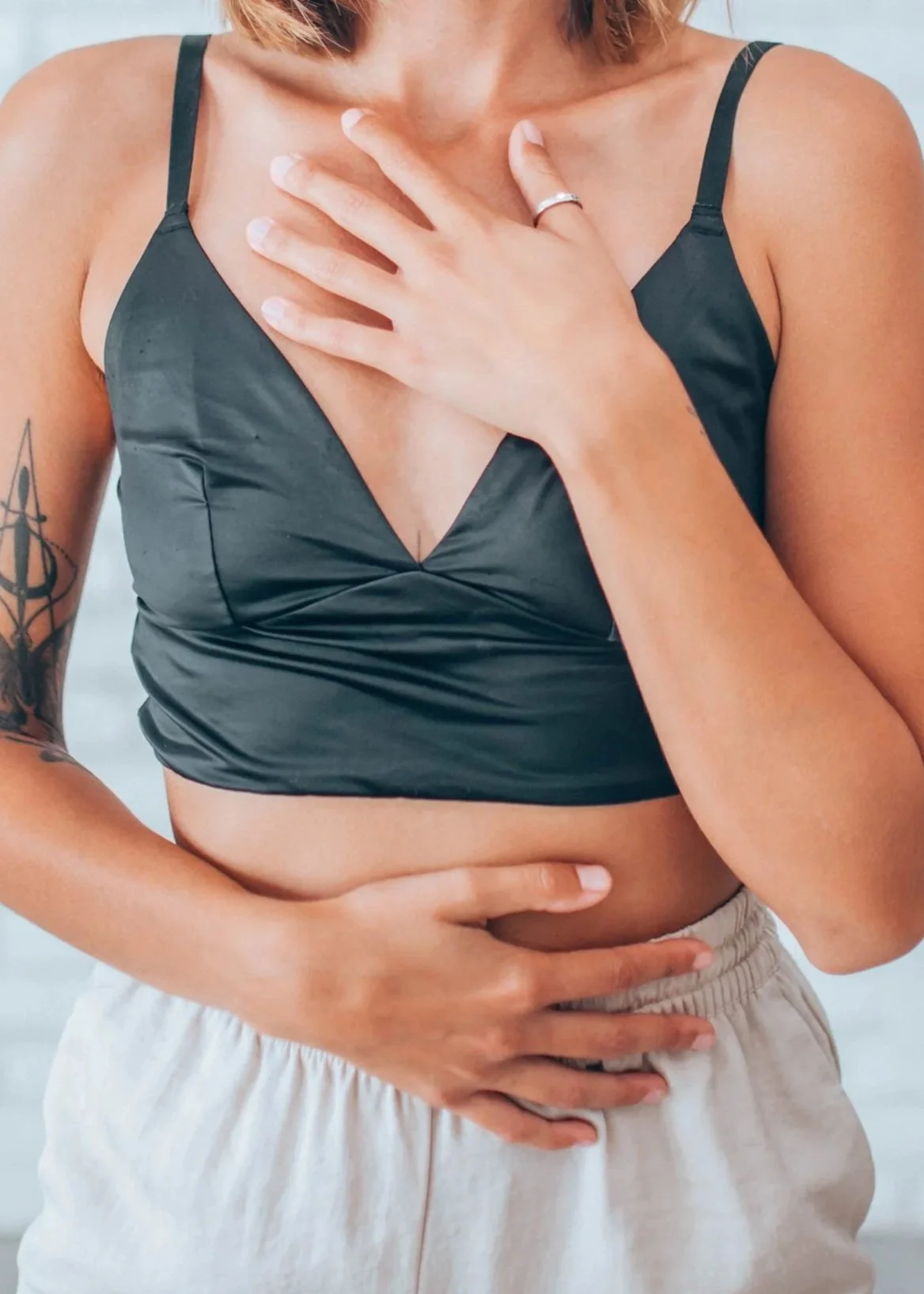 A woman touching her chest with her right hand and her stomach with her left hand, wearing a black satin crop top and beige pants, with a tattoo on her left arm.
