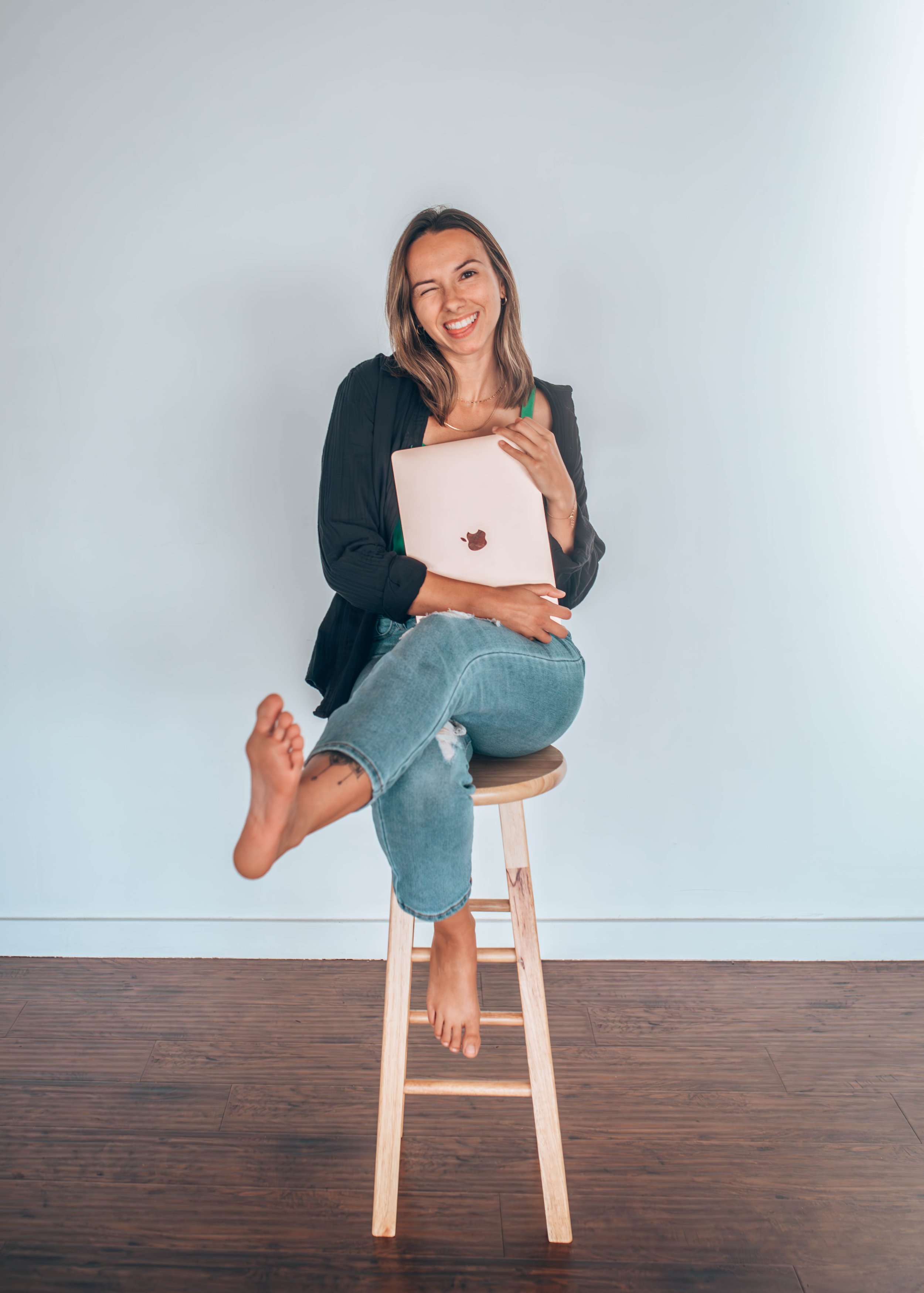 A woman sitting on a wooden stool, holding a laptop, smiling and winking, with an outstretched arm and a barefoot foot in the foreground, against a plain white wall.