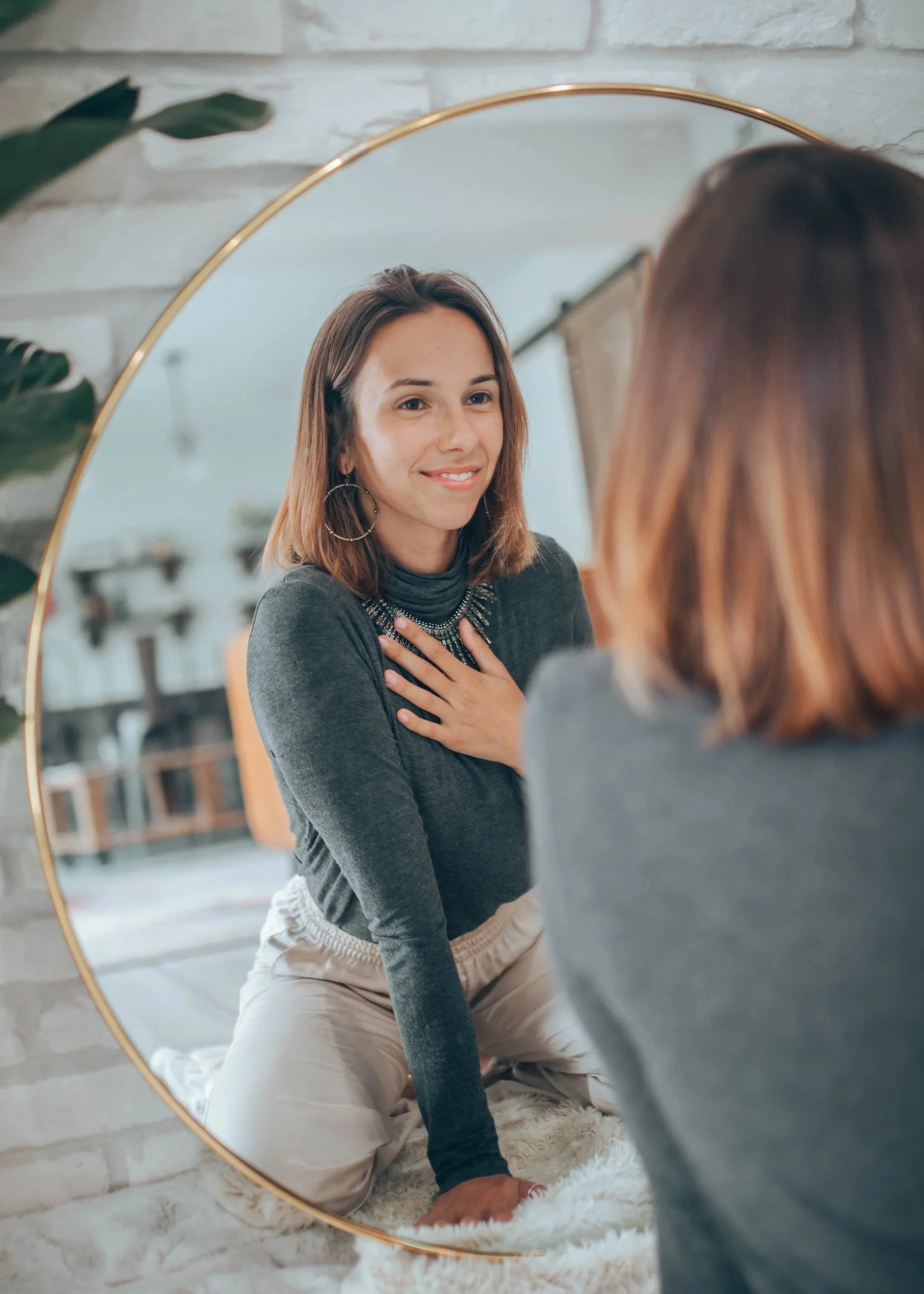 Woman with shoulder-length brown hair looking into a round mirror, smiling, wearing large hoop earrings, dark long-sleeve shirt, sitting on a fluffy white rug near a white brick wall and plant leaves.