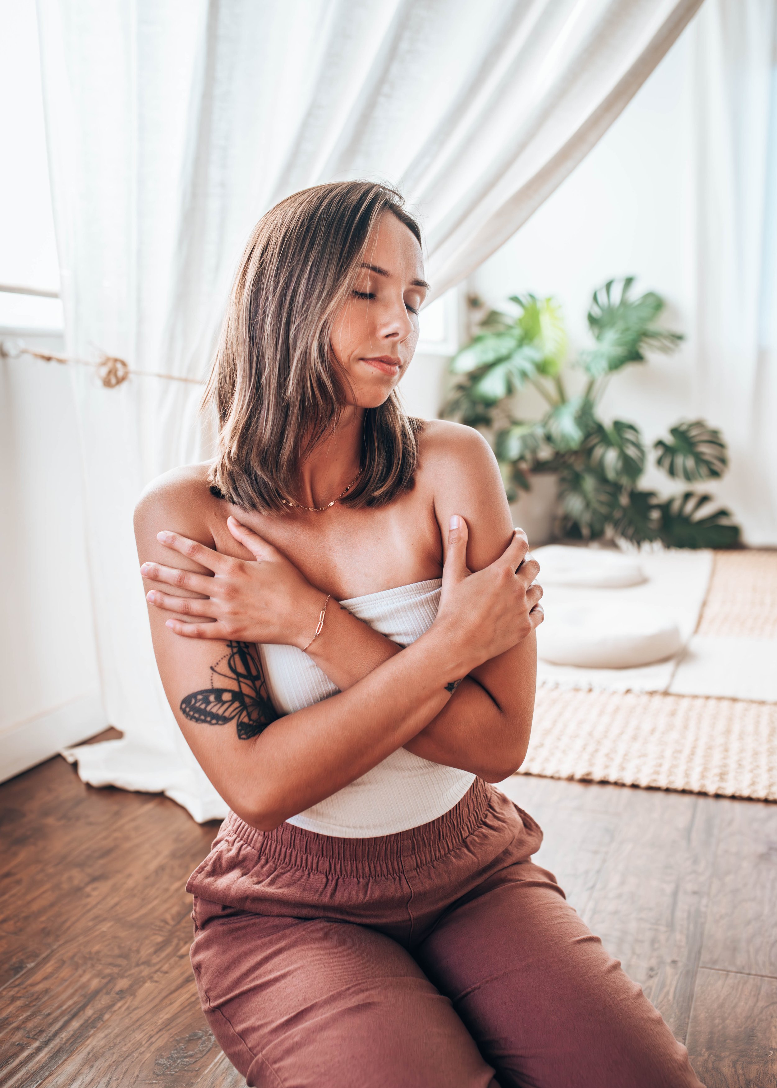 A woman with shoulder-length brown hair, wearing a white strapless top, tan pants, and a delicate necklace, sitting indoors with her eyes closed and arms crossed over her chest. There is a large green plant, a rug, and a white basket in the background, with sunlight coming through a window covered by light curtains.
