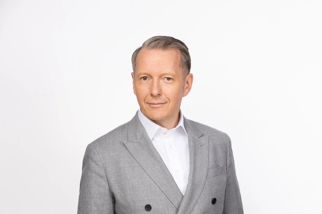 Ein Mann in einem grauen Anzug und weißem Hemd vor einem weißen Hintergrund.