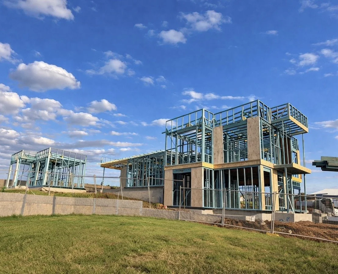 Under construction building with steel framing and large windows, set on a grassy hill under a blue sky with scattered clouds.