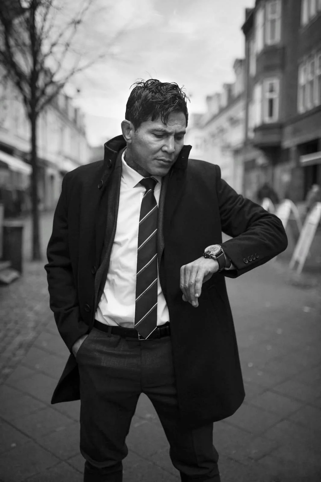 A man in a business suit and tie in a street, looking down at his watch, wearing a dark overcoat.