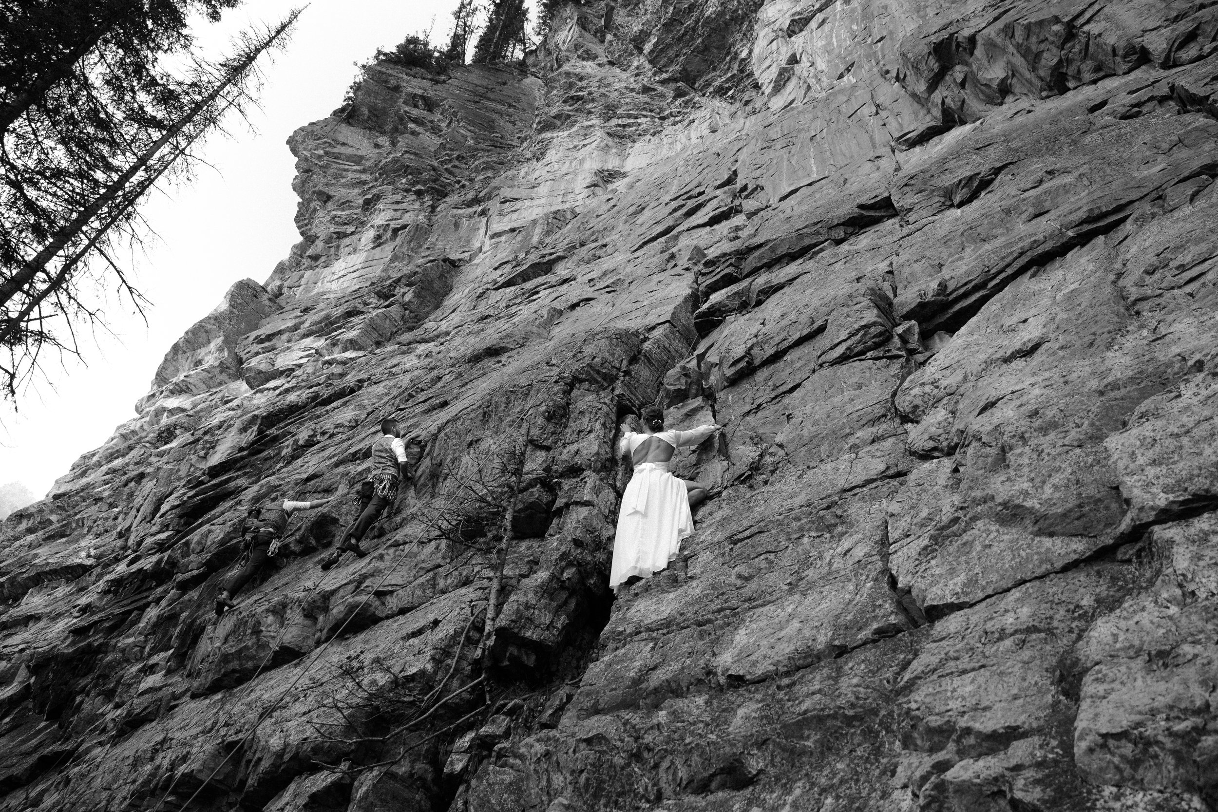 Black and white photo of a woman in a white dress climbing a rugged rock face, with two men assisting further down.