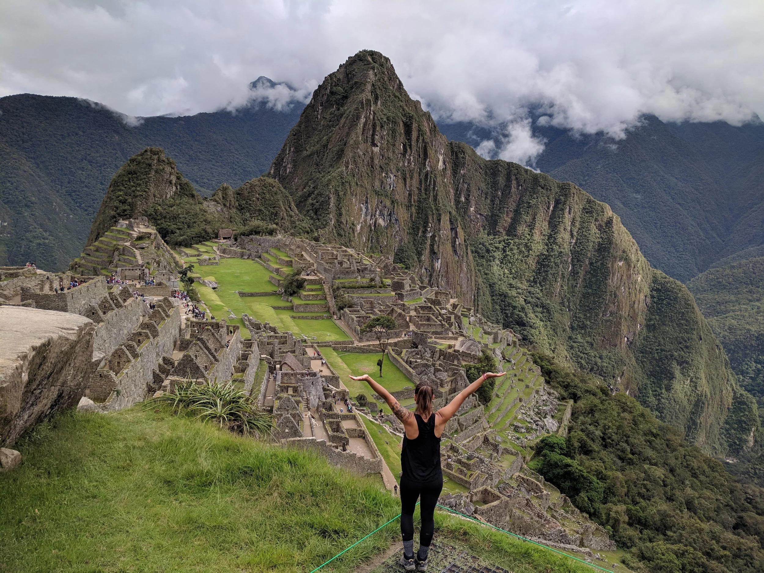 A woman with outstretched arms stands on green grass overlooking Machu Picchu with ancient stone ruins and green mountains with clouds in the background.