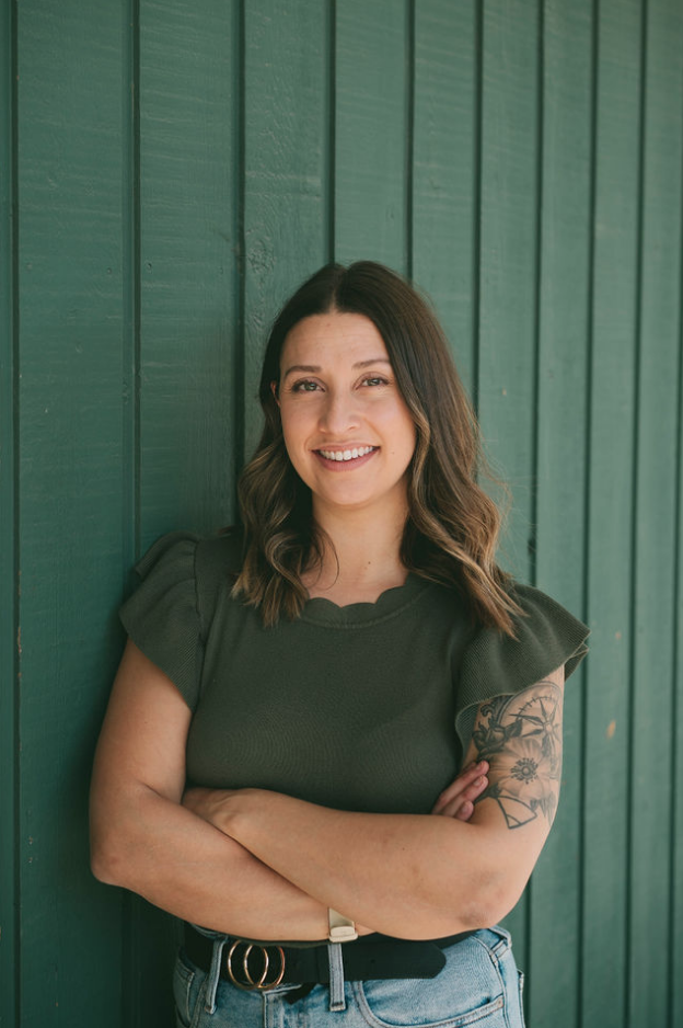 A woman with wavy brown hair, smiling, standing against a green wooden wall, wearing a dark green top and jeans with her arms crossed.