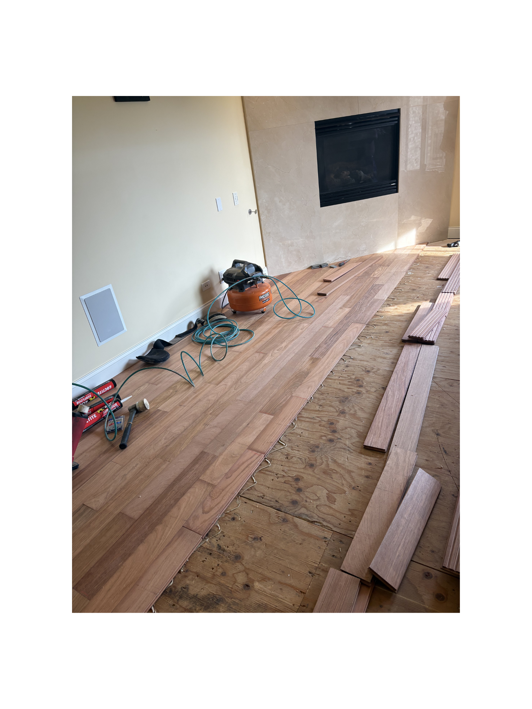 Installing hardwood flooring in a living room with a stone fireplace, partially finished floor, and construction tools.