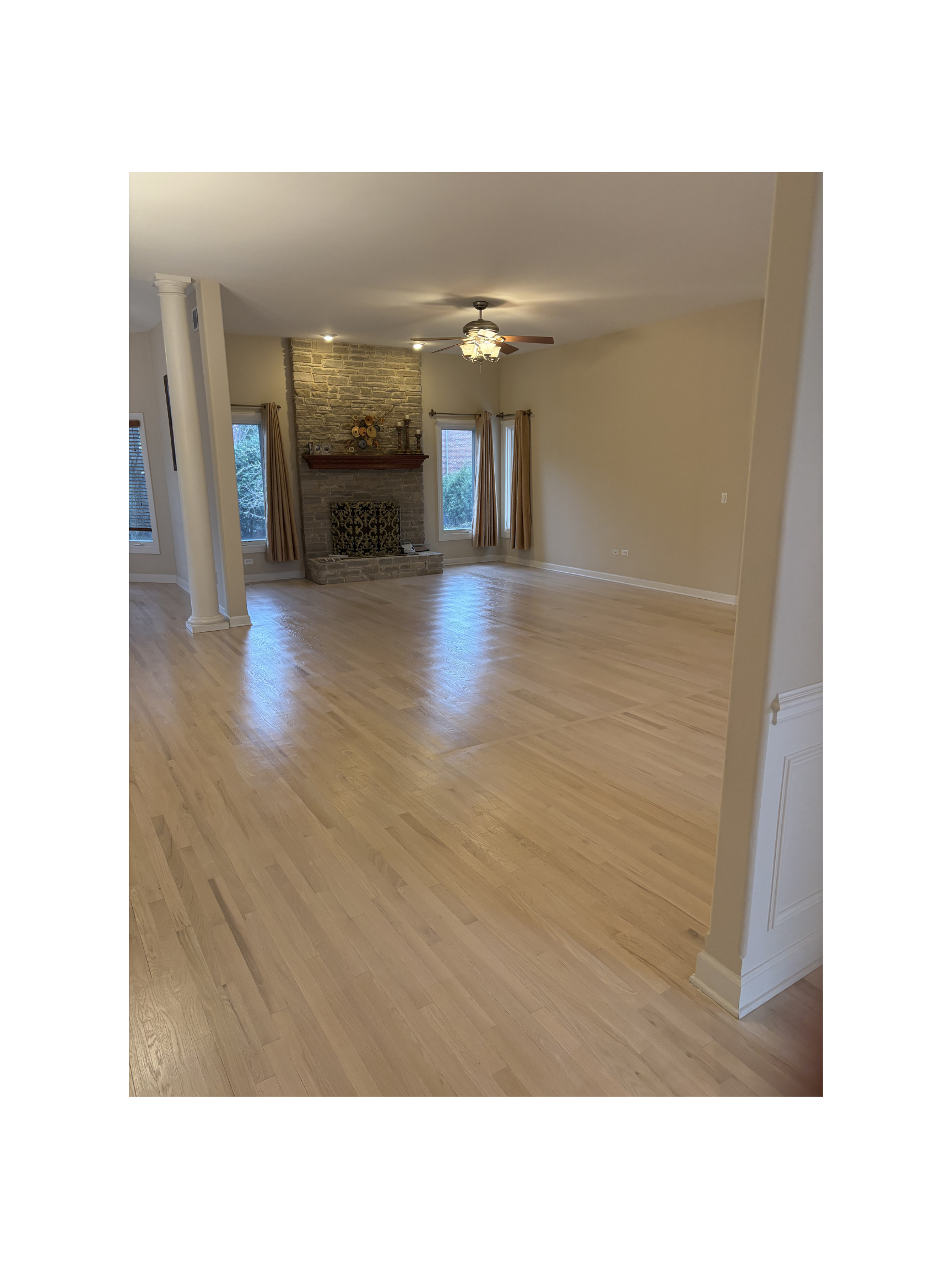 Empty living room with hardwood floors, brick fireplace, ceiling fan, and large windows with beige curtains.