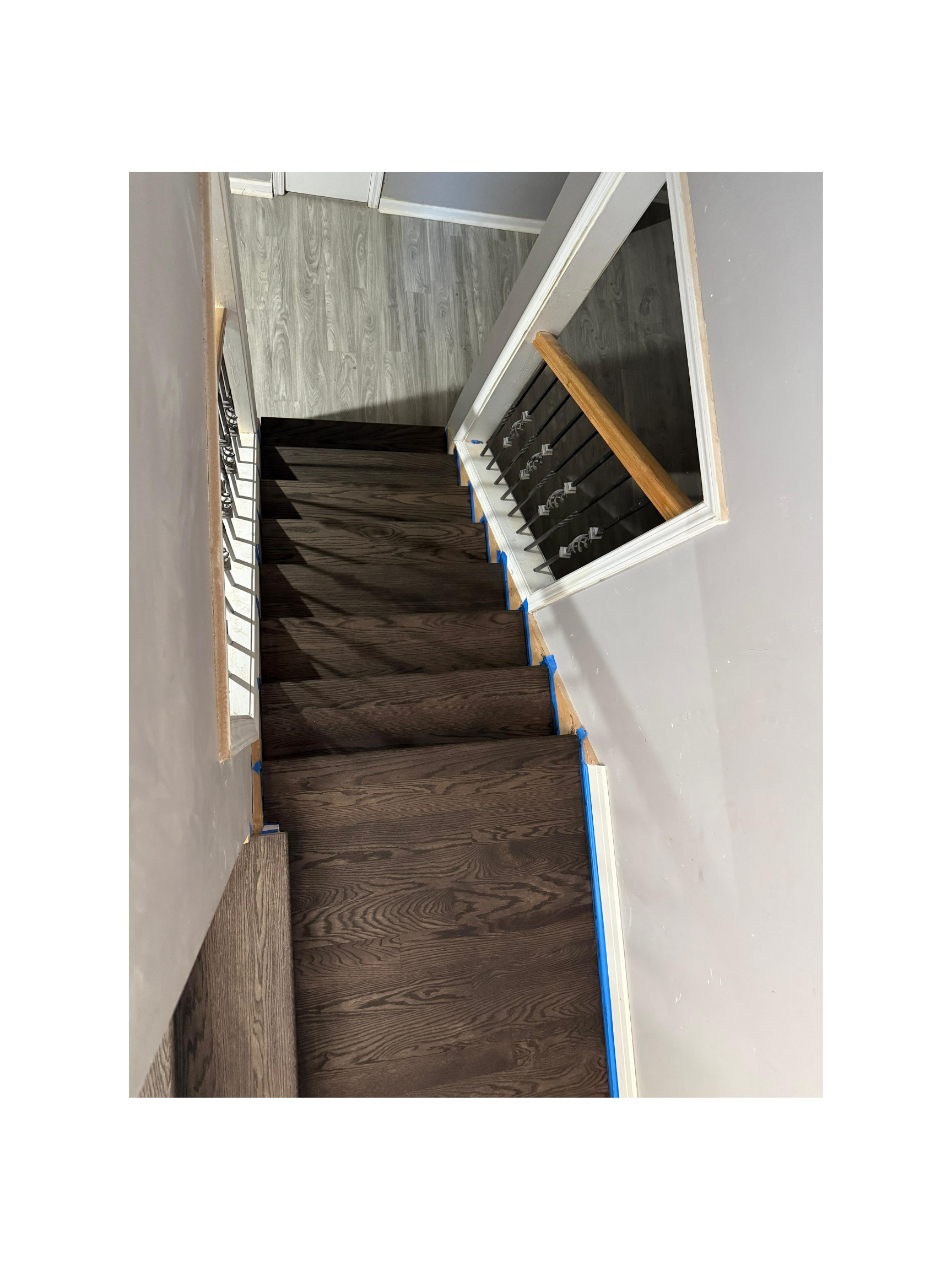 Interior view of a staircase with dark wood treads and no risers, with blue painter's tape along the edges, leading down to a lighter wood floor, with a black metal railing and a wooden handrail on the right side.