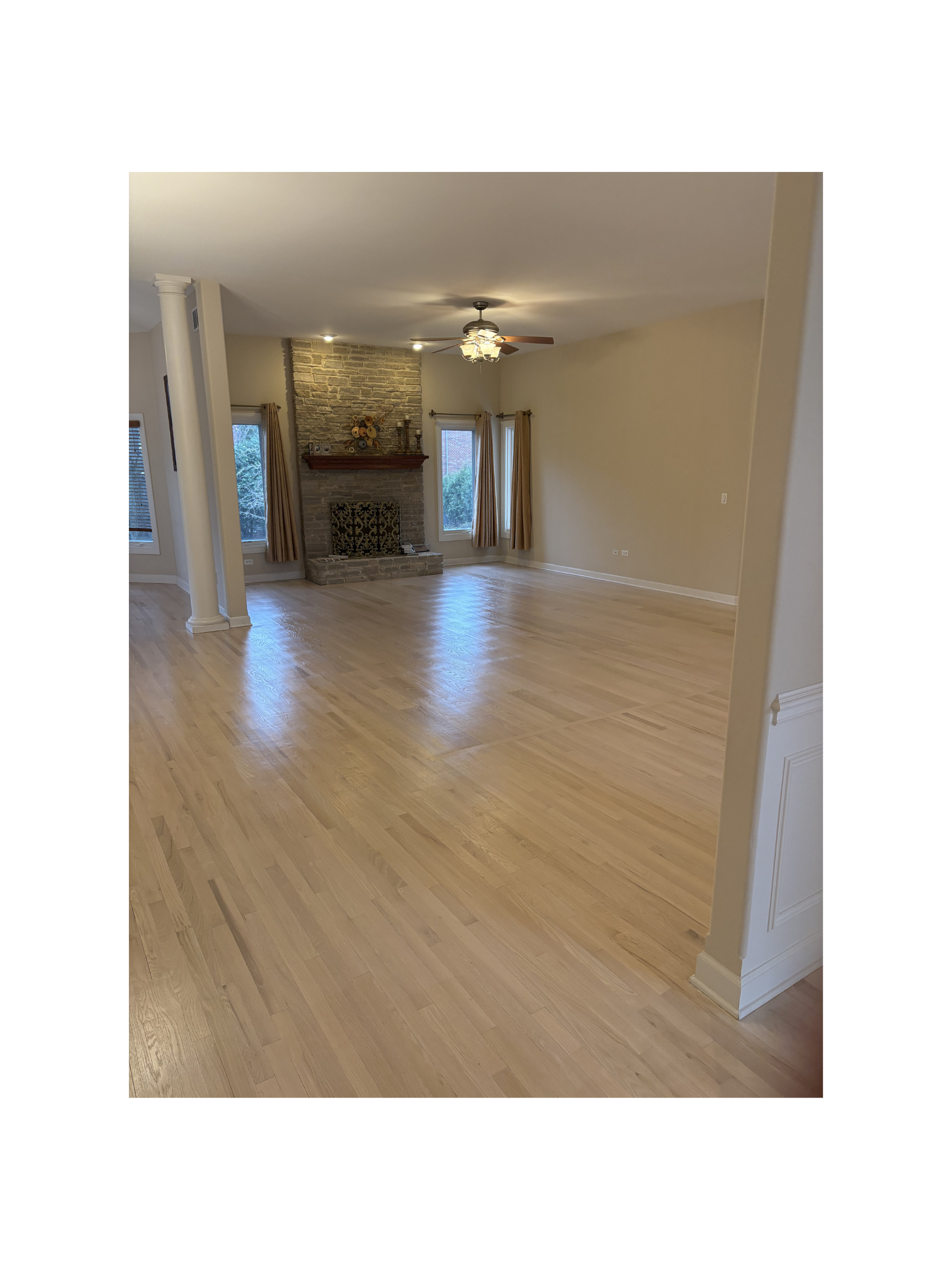 Empty living room with wooden floors, a brick fireplace, and large windows with curtains.