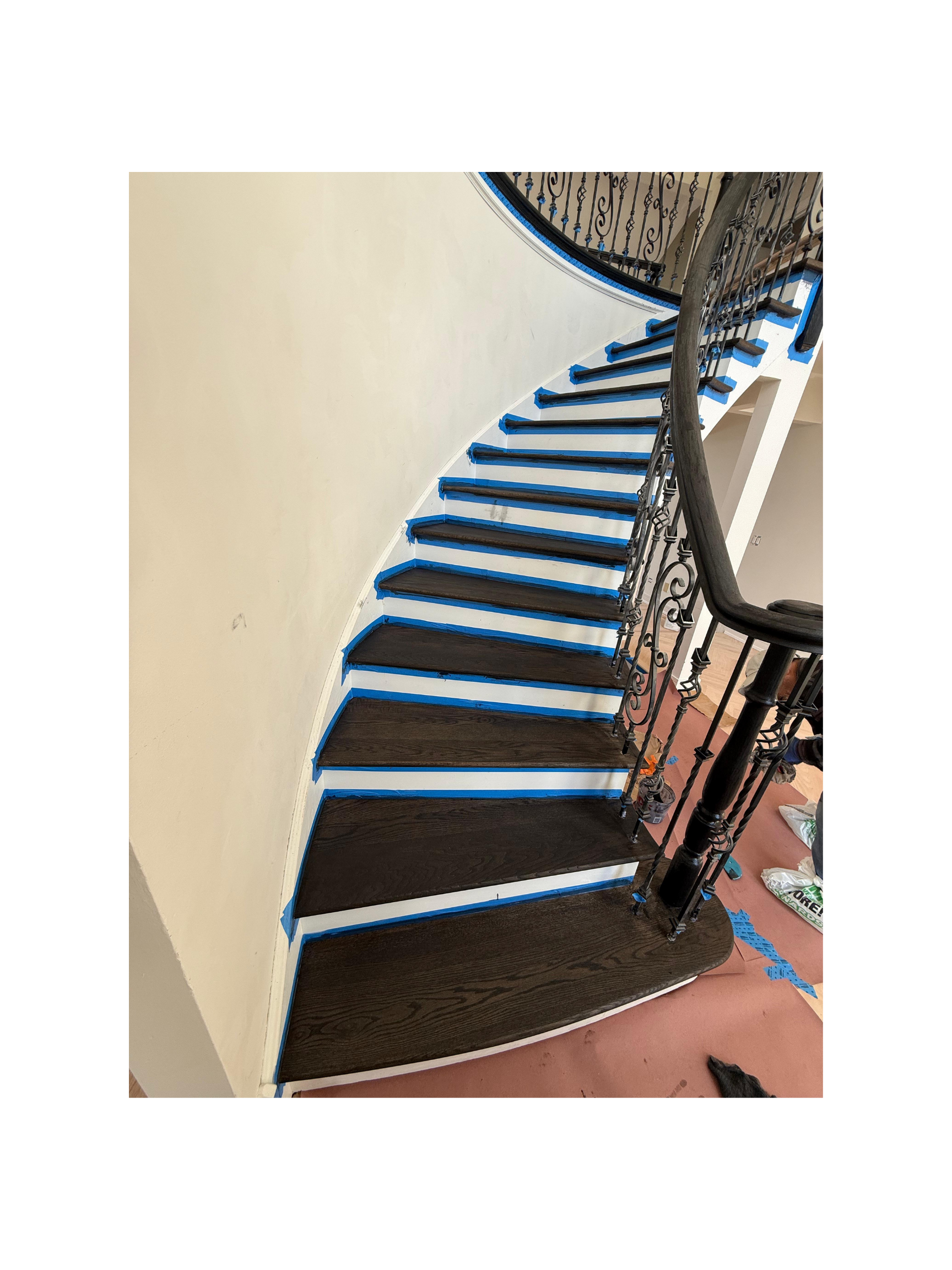 Spiral staircase with dark wooden steps, ornate metal railing, and blue painter's tape along the edges, with construction supplies on the floor nearby.