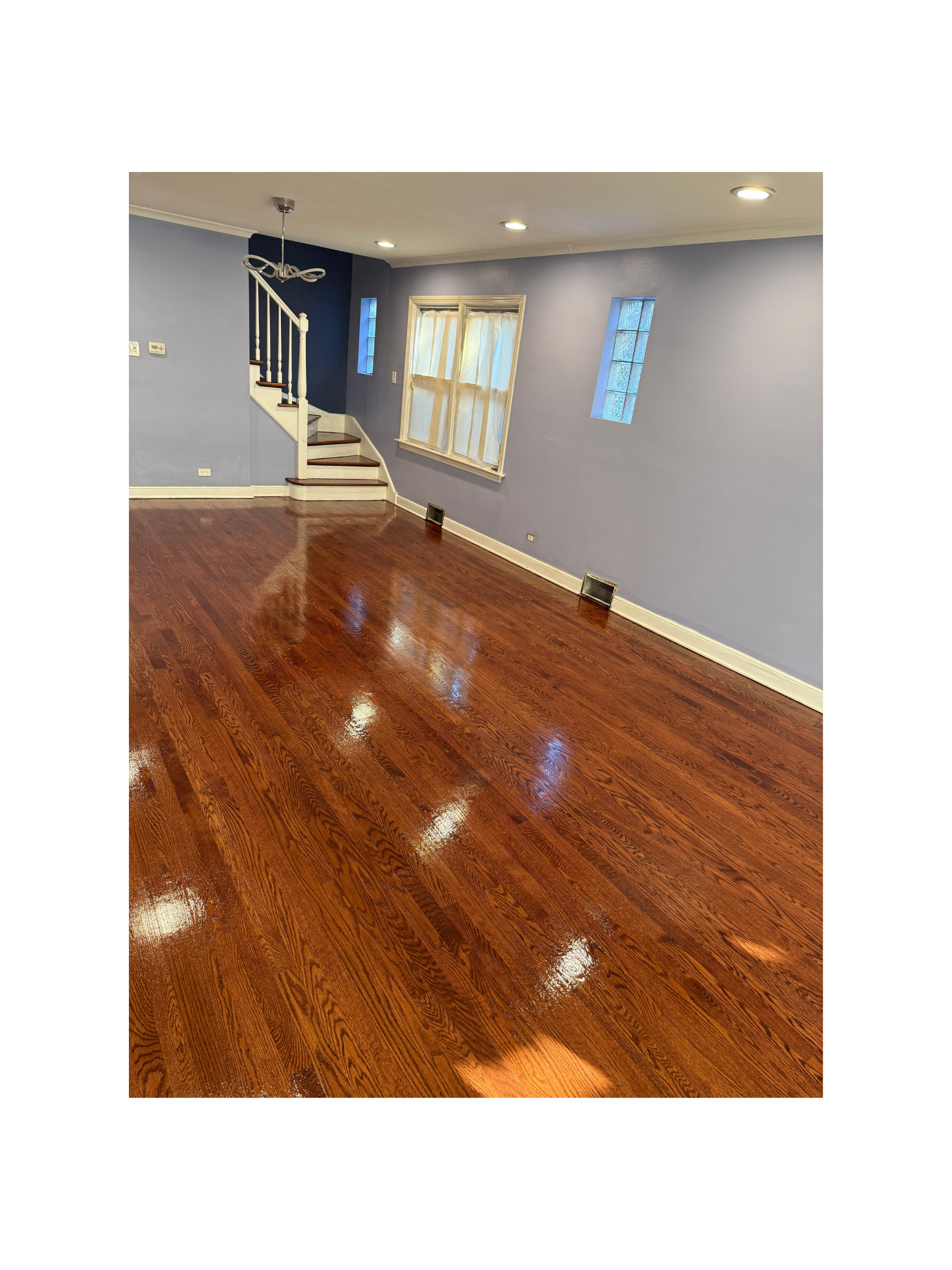 Empty living room with wooden floor, light blue walls, a few small windows, a set of stairs with a white railing, and ceiling lights.