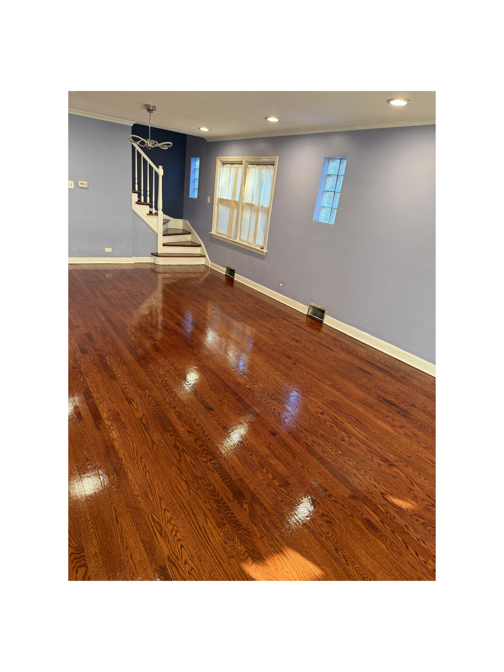 Empty living room with polished hardwood floors, blue walls, a staircase with white railings, and two narrow windows.
