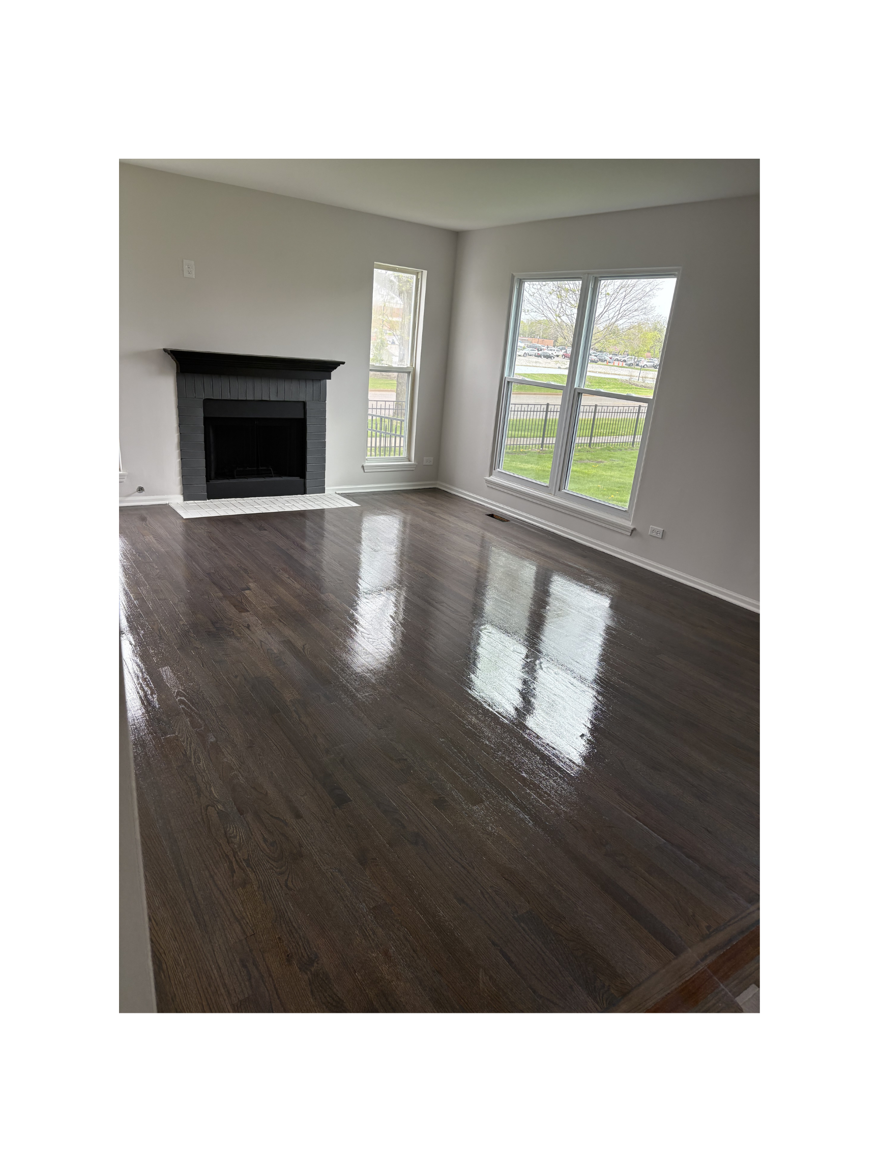 Empty living room with shiny hardwood floors, a black fireplace, and large windows showing a grassy outdoor area.