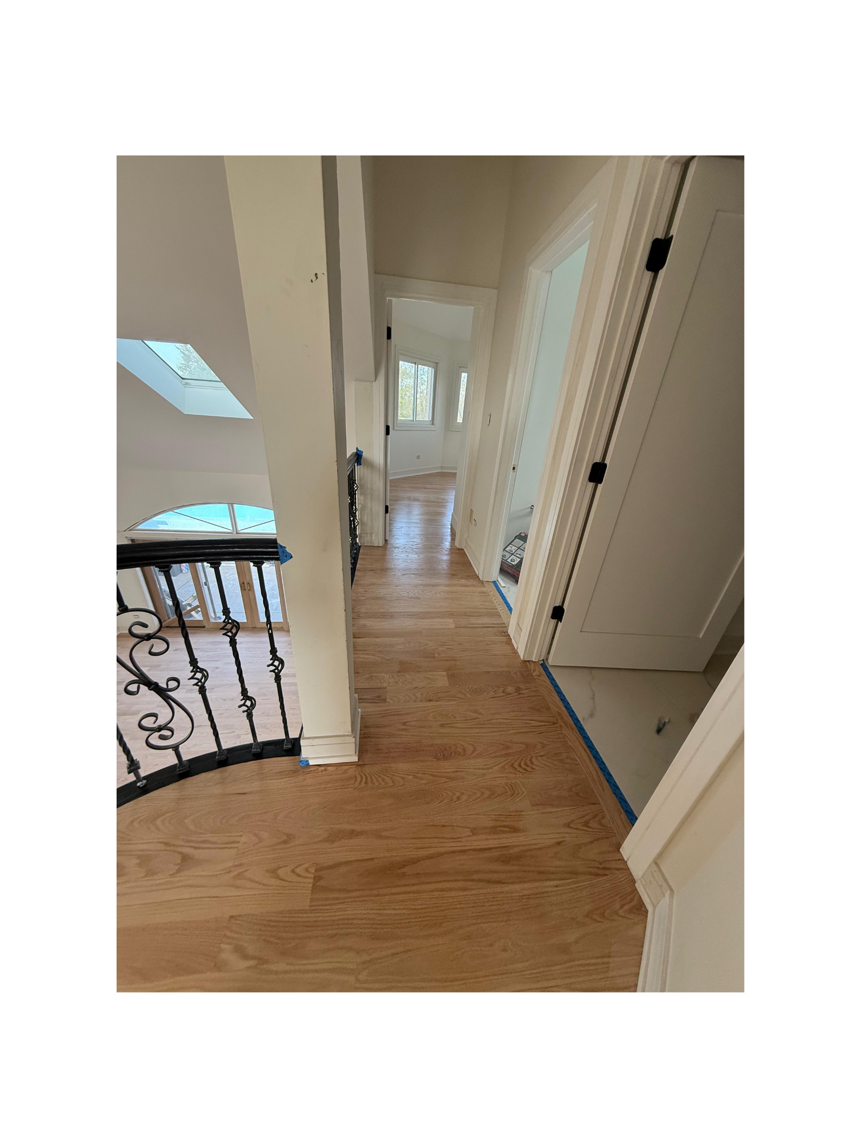 Interior view of a freshly renovated house hallway with wooden flooring, white walls, multiple doorways, and a staircase with a black wrought-iron railing.