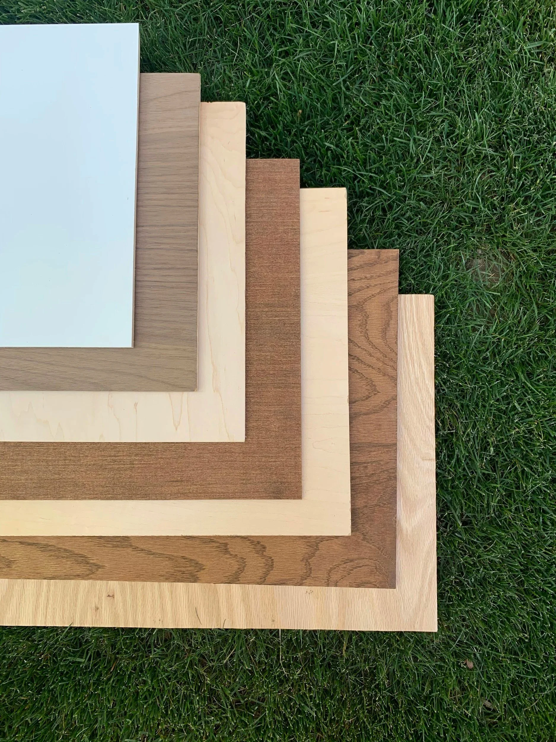 Stacked wooden boards of various colors and textures, placed on green grass, with the largest white board on top.